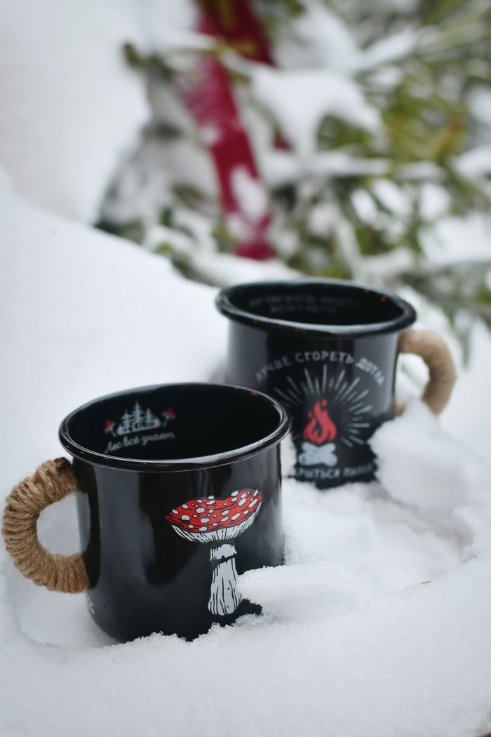 Two black mugs with unique designs resting on fresh snow, capturing winter aesthetics.